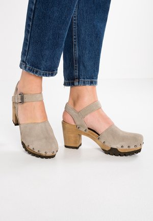 Beige suède klompen met hakjes, enkelriempjes en houten plateauzolen, gedragen met cropped blauwe jeans op een witte achtergrond.