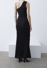 Robe maxi noire avec une bretelle et un corsage ajusté, fabriquée en tissu lisse. Fente à l'arrière, associée à des talons ouverts.