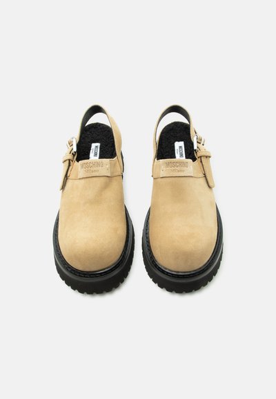 Μπεζ σουέντ κλειστά υπόδημα με μαύρες παχιές καουτσούκ σόλες, ρυθμιζόμενα πλευρικά λουριά και μαύρη εσωτερική επένδυση με το λογότυπο Moschino Milano.