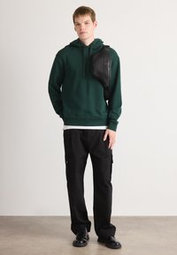 Zielona bluza z kapturem, czarne spodnie cargo z bocznymi kieszeniami, czarna torba na pasek i czarne buty. Prosty design, miękka tekstura, luźny krój.