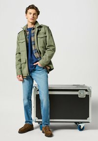 Olivgrön militärjacka över en blå skjorta, med rutig flanell detaljer. Blå jeans och bruna mockaskor. Förvaringsfodral bredvid.
