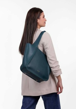 Woman with long brown hair wearing a beige blazer and blue jeans carries a large dark teal leather shoulder bag.