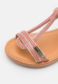 Sandale en cuir rose avec des lanières entrecroisées, des accents dorés et un semelle beige. Comprend une étiquette en métal sur le côté et des détails cousus.