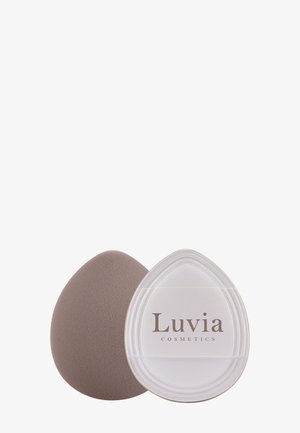 Esponja de maquillaje de color topo, forma ovalada con textura suave, junto a un envase transparente grabado con la etiqueta "Luvia Cosmetics".