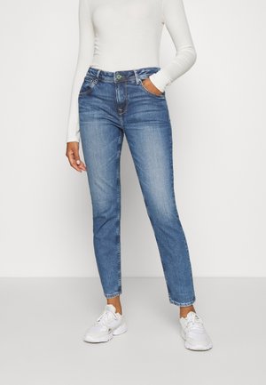 Frau trägt blaue Slim-Fit-Jeans, weißes langärmeliges Oberteil und weiße Sneakers, steht mit einer Hand in der Tasche.