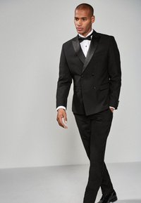 Sort dobbeltradet smokingjakke med satinrevers, hvid skjorte, sort butterfly og matchende bukser, båret med polerede sorte sko.