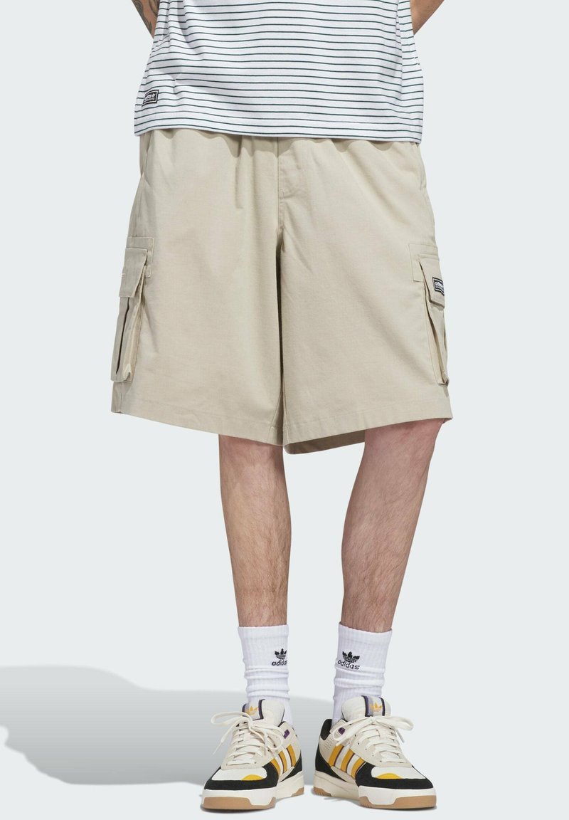Hombre con pantalones cortos cargo beige, calcetines Adidas blancos hasta la mitad de la pantorrilla, zapatillas de estilo retro con rayas amarillas y negras, de pie sobre un fondo liso.