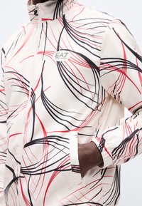 Λευκό μπουφάν με κόκκινες και μαύρες αφηρημένες γραμμές, φερμουάρ μπροστά, λογότυπο στο στήθος, φορεμένο από άτομο με το χέρι στην τσέπη.