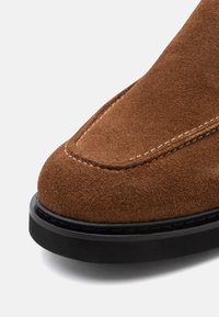 Brown suede shoe featuring a rounded toe, contrasting stitching, and a black rubber sole. The texture is soft and velvety.
