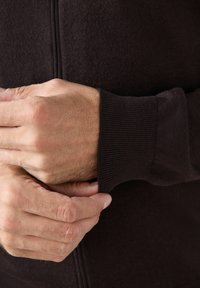 Ręce dostosowujące mankiet ciemno-brązowego swetra z dzianiny, ukazujące fakturę i detale tkaniny.