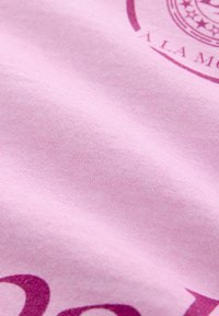 Gaiši rozā audums ar gludu tekstūru, kas rotāts ar tumši rozā druku, kurā ir teksts un apļveida grafiskais dizains.