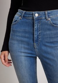 Jeans de mezclilla azules de tiro alto con un ligero desgaste, bolsillos frontales y cierre de botón plateado. Textura suave y diseño ajustado.