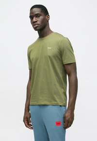 Olivgrünes Baumwoll-T-Shirt mit kurzen Ärmeln, das ein weißes Logo auf der linken Brust zeigt. Glatte Textur, Rundhalsausschnitt.