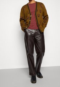 Pantalones de cuero marrón combinados con un cárdigan mostaza que presenta patrones florales oscuros y botones, y una camiseta burdeos debajo.