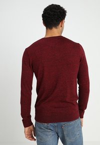 Punainen neulepaita pitkillä hihoilla, pyöreällä kaula-aukolle ja ribbireunuksella. Varpustelevalle sinisten farkkujen päällä, esittää tekstuurillista kangaskuvioita.