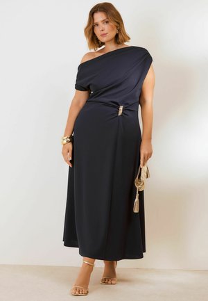 Woman wearing a one-shoulder black midi dress with gold buckle, gold bracelet, holding a gold clutch, and wearing gold sandals.