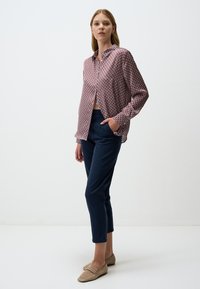 Roter und blauer gemusterter Hemd mit Knöpfen, lange Ärmel, kombiniert mit dunkelblauen, enganliegenden Hosen. Hellbraune Slipper. Minimalistischer Hintergrund.