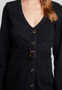 Svart denimklänning med V-ringning, långa ärmar, midjebälte och bruna knappar framtill. Texturerad tyg med en strukturerad passform.