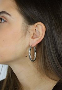 Teksturowane srebrne kolczyki-koła z przekręconym wzorem i subtelnym ornamentem, prezentowane na uchu modelki na tle długich brązowych włosów.