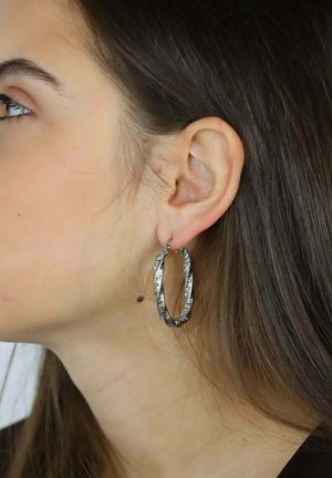 Teksturowane srebrne kolczyki-koła z przekręconym wzorem i subtelnym ornamentem, prezentowane na uchu modelki na tle długich brązowych włosów.