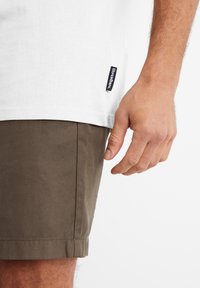 Hvid bomulds t-shirt med korte ærmer, med et lille sort mærke i kanten. Olivengrønne shorts med lige snit og kant.