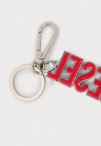 Piros akril kulcstartó, amelyen a "SHE" felirat látható, fényes ezüst szegéllyel és fémkapoccsal, kör alakú kulcskarika rögzítve.