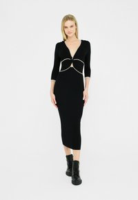La modella si erge con fiducia in un vestito nero aderente con scollo profondo e stivali neri con lacci. Sfondo bianco semplice.