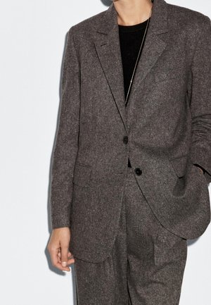 Person wearing a dark gray wool suit with two buttons and flap pockets, paired with a black top and a thin gold necklace.