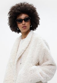 Λευκό φουντωτό μπουφάν από shearling με γιακά σαλονιού, φορεμένο πάνω από ένα ελαφρύ τοπ, συνδυασμένο με μαύρα γυαλιά ηλίου τύπου cat-eye και ασημένια σκουλαρίκια κρίκους.