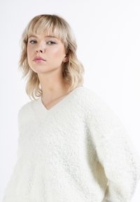 Pulover alb, pufos, supradimensionat, cu un decolteu adânc în V și tricotaj texturat. Material moale și confortabil, care reflectă ușor lumina de pe suprafață.