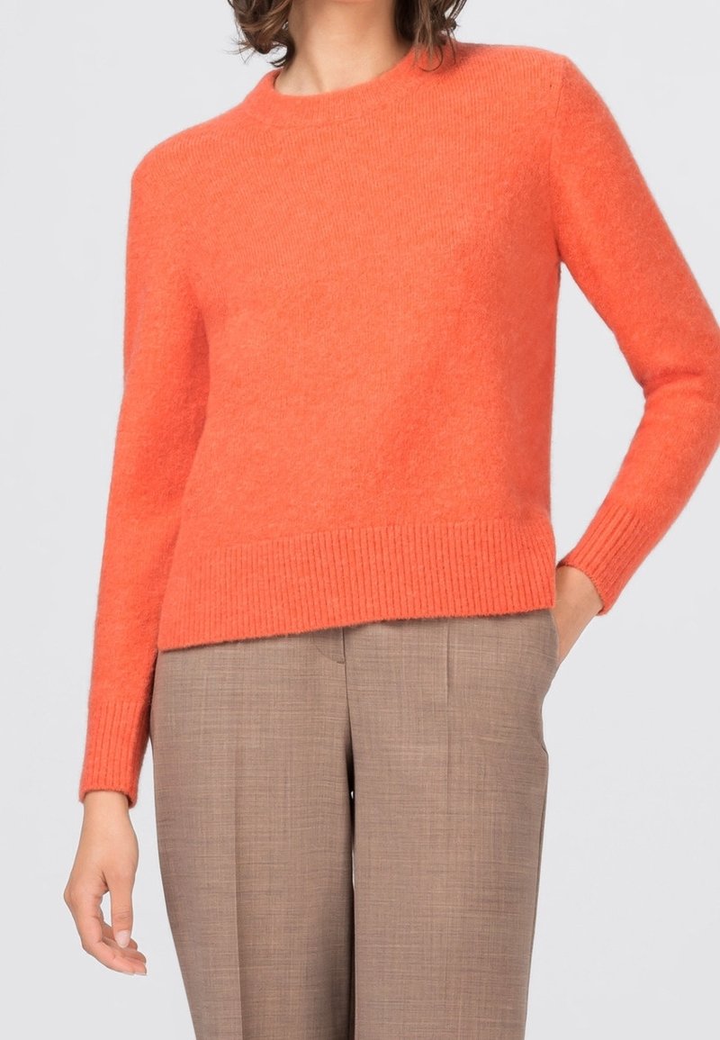 Personne portant un pull à manches longues orange vif et un pantalon beige sur mesure, avec une main dans la poche, sur un fond clair uni.