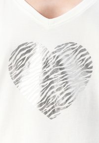 T-shirt en coton blanc avec un motif de cœur rayé argenté au centre, à col en V et texture douce.