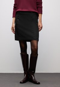 Mini-jupe noire avec des boutons sur le devant, associ�ée à des bottes brillantes marron hauteur genou et des collants noirs. Le matériau semble lisse et structuré.