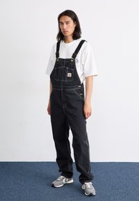 Sorte denim overaller med justerbare stropper, frontlomme, sølvfarget maskinvare og hvite sting; kombinert med en hvit t-skjorte og grå joggesko.