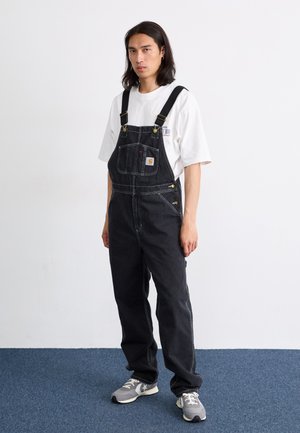 Black denim overalls with adjustable straps, front pocket, silver hardware, and white stitching; paired with a white t-shirt and gray sneakers.