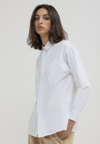 Junge Frau mit schulterlangem dunklem Haar, die ein weißes Langarmhemd und beige Hosen trägt, steht vor einem schlichten Hintergrund.