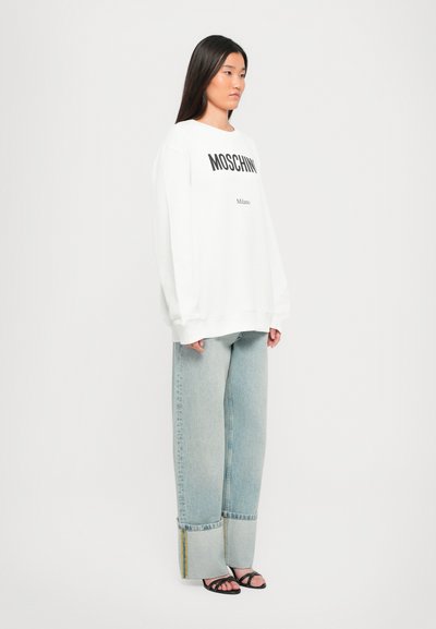 Vit oversized sweatshirt med "MOSCHINO" och "Milano" tryckt i svart. Bärs med ljusblå vida jeans och svarta sandaletter.