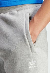 Grå bomullsoveraller med sidofickor, med en liten vit Adidas-logotyp. Midjebandet är elastiskt med en slät yta.