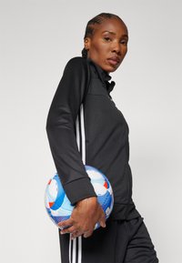 Veste de sport zippée noire avec des rayures blanches, portée avec un pantalon assorti. La personne tient un ballon de soccer multicolore avec des motifs bleus, rouges et blancs.