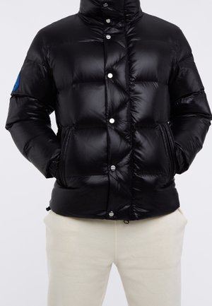 Black shiny puffer jacket with snap buttons and zippered pockets worn over light beige pants, hands in pockets, standing against plain background.
