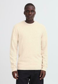 Light beige knit sweater with a rounded neckline and ribbed cuffs, made from a soft material, paired with dark trousers.