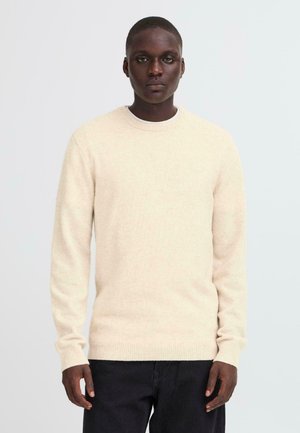 Maglione lavorato a maglia di un beige chiaro con scollatura tonda e polsi a coste, realizzato in un materiale morbido, abbinato a pantaloni scuri.