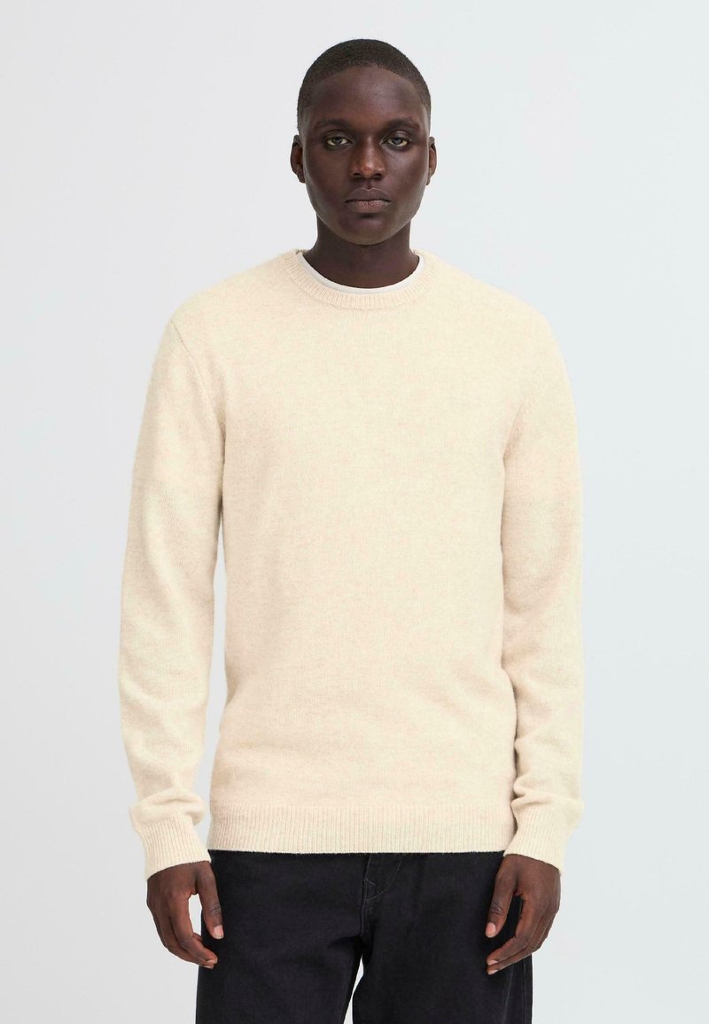 Light beige knit sweater with a rounded neckline and ribbed cuffs, made from a soft material, paired with dark trousers.