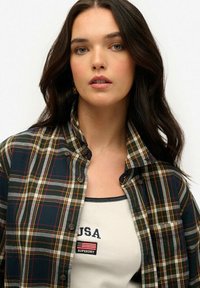 Chemise à carreaux boutonnée dans des tons de vert foncé, rouge et jaune ; tissu léger avec un col classique. Débardeur blanc avec un logo brodé visible en dessous.