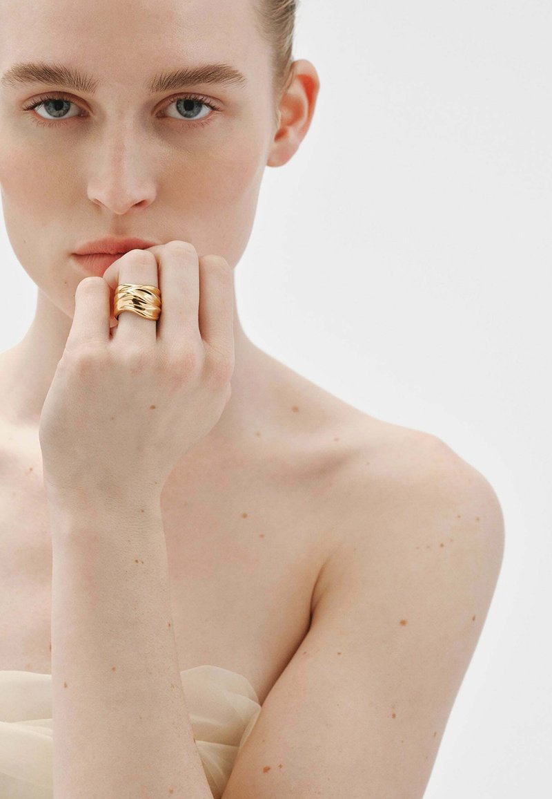 Anillo de color dorado con un diseño ondulado, llevado en una mano de piel clara. El fondo es blanco, con una iluminación suave y natural que resalta los detalles.