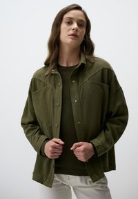 Olijfgroene oversized jas met drukknoopsluiting, borstzakken en een gladde textuur, gedragen over een donkergroen sweater en witte broek.