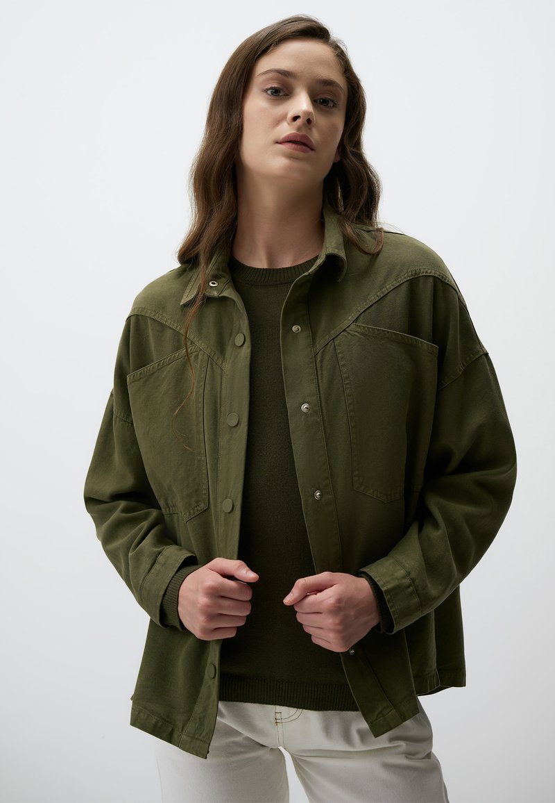Olijfgroene oversized jas met drukknoopsluiting, borstzakken en een gladde textuur, gedragen over een donkergroen sweater en witte broek.
