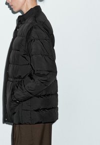 Woman in profile wearing a black puffer jacket with hands in pockets against a plain light background.