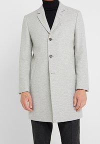 Abrigo de lana gris con un corte entallado, que presenta solapa con muesca, dos botones marrones, bolsillos laterales y textura suave. Llevado sobre un suéter de cuello alto negro.