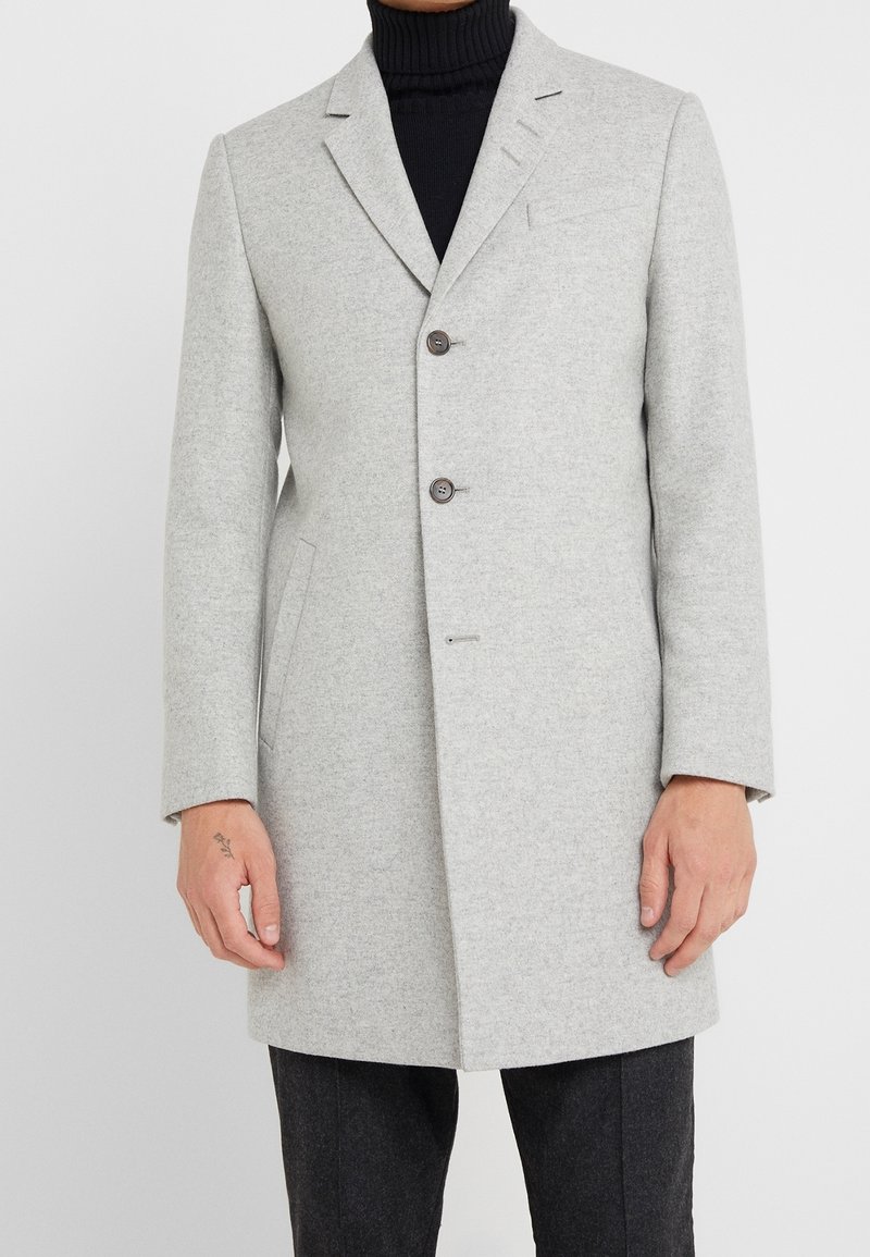 Abrigo de lana gris con un corte entallado, que presenta solapa con muesca, dos botones marrones, bolsillos laterales y textura suave. Llevado sobre un suéter de cuello alto negro.